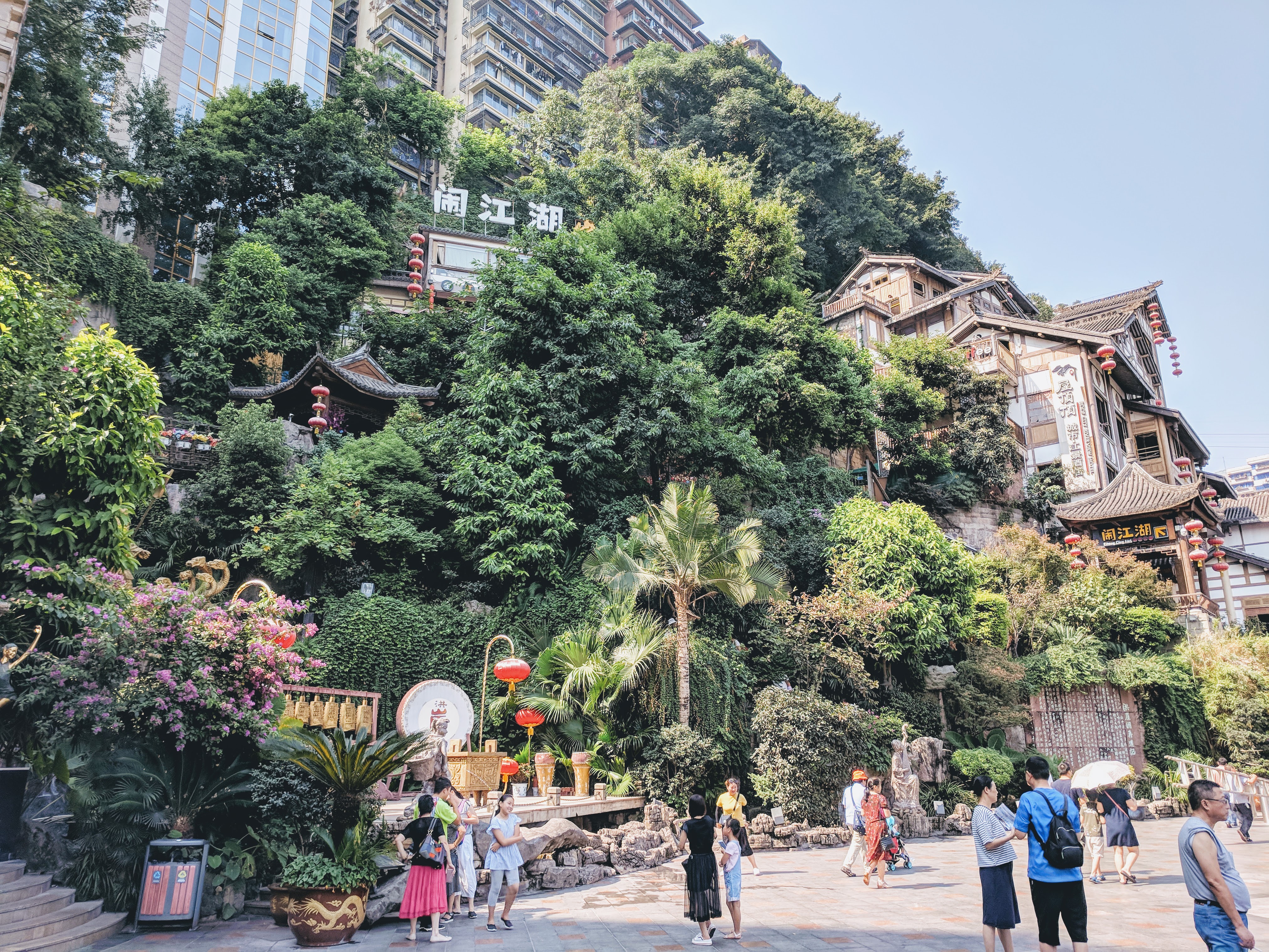 People standing before several old-school chinese buildings, green foliage everywhere.